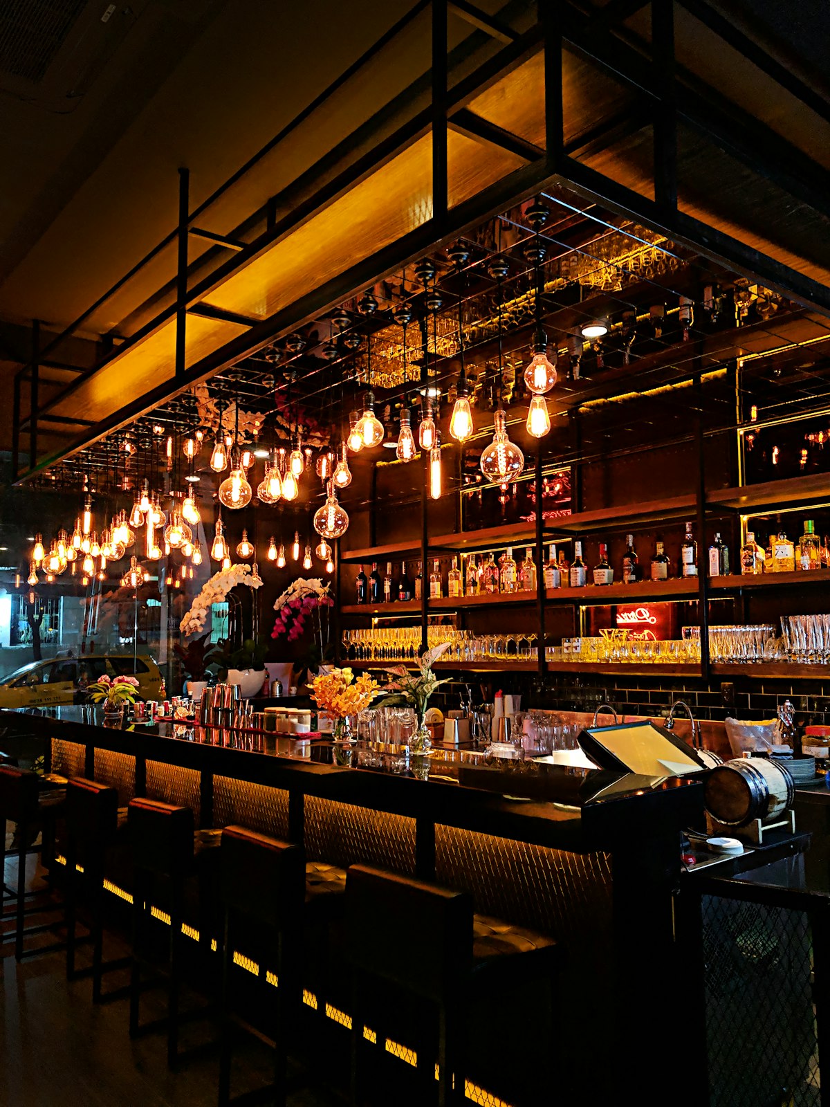 A long dark bar counter lit by hanging filament bulbs, with spirit bottles and empty bar stools.