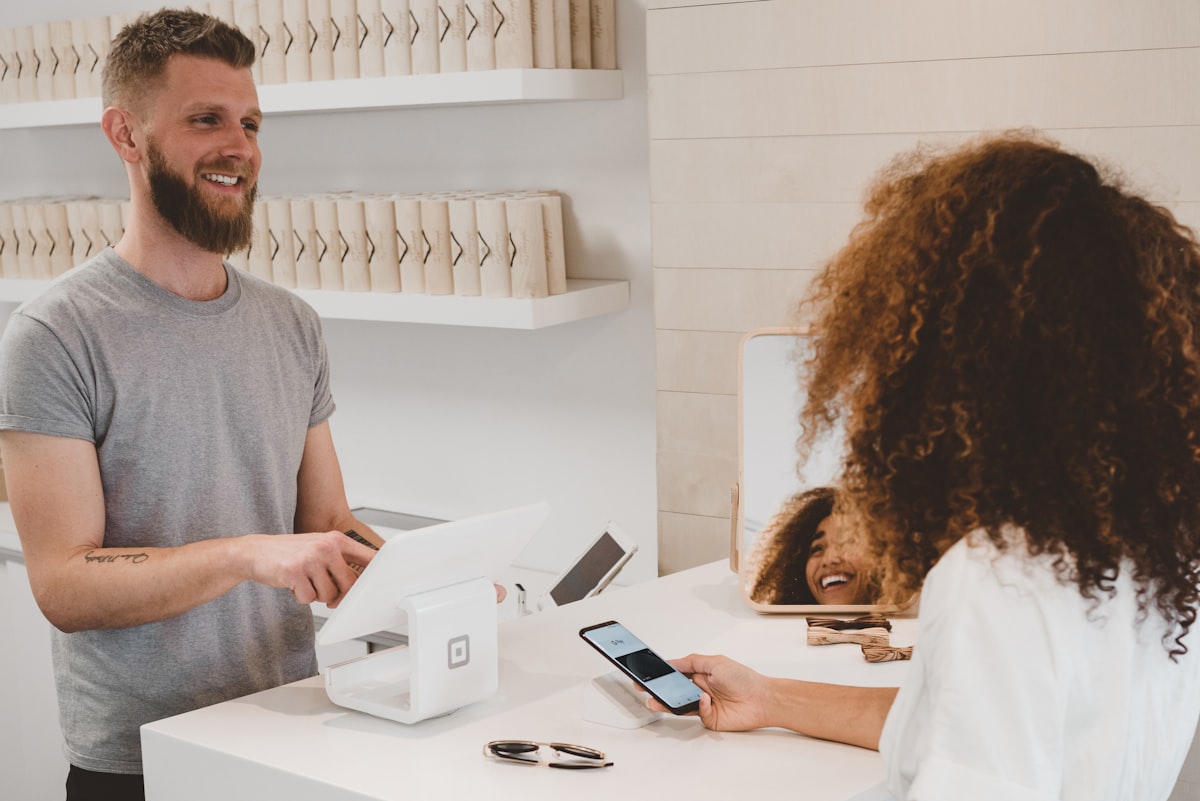 A bearded shop assistant serves a customer paying by phone at a white tablet till.