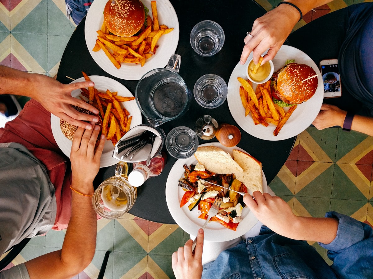 Overhead view of several people sharing burgers, chips and drinks around a small round table.