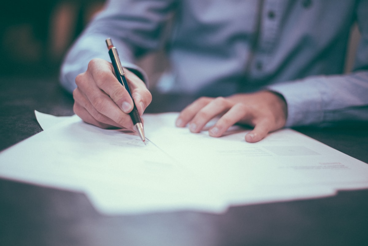 A person in a pale shirt signs a stack of printed documents with a pen.