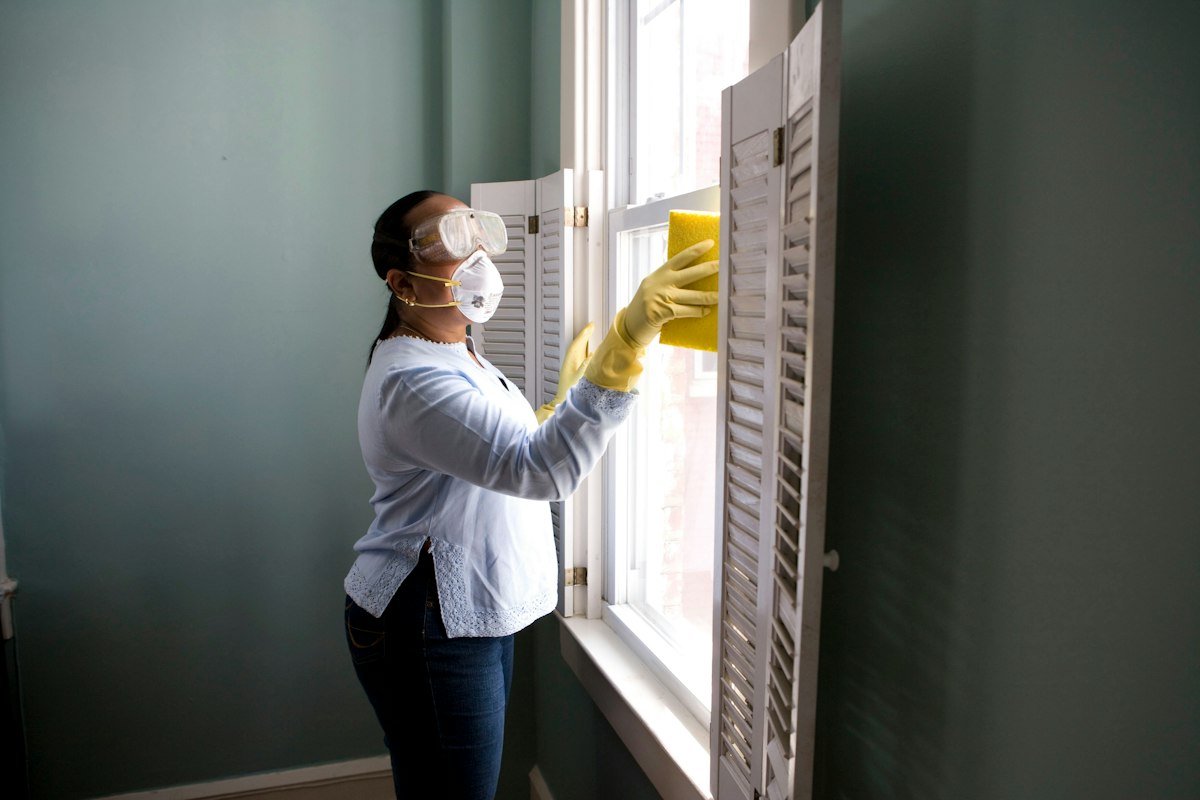 A woman in safety goggles, face mask and yellow rubber gloves cleans a window with a sponge.