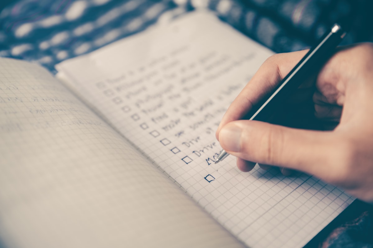 A hand with a pen draws tick boxes down a checklist in a squared paper notebook.