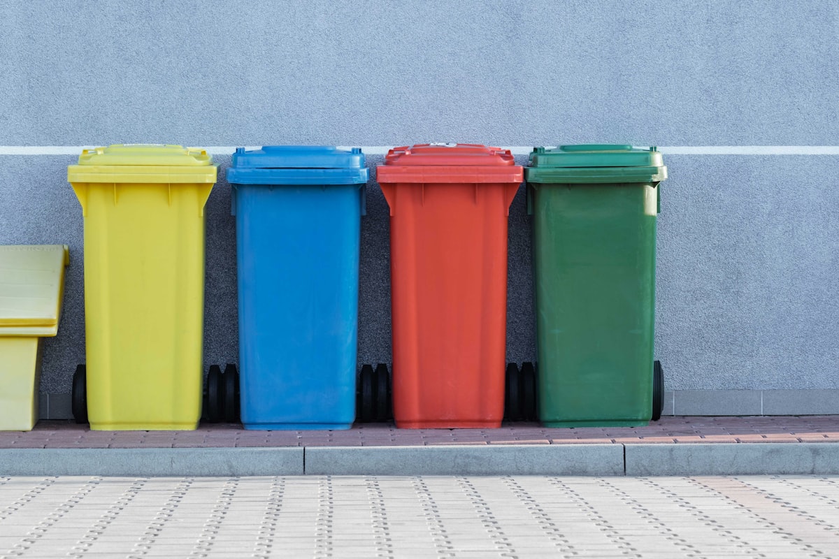 Yellow, blue, red and green wheelie bins lined up against a grey wall on a pavement.