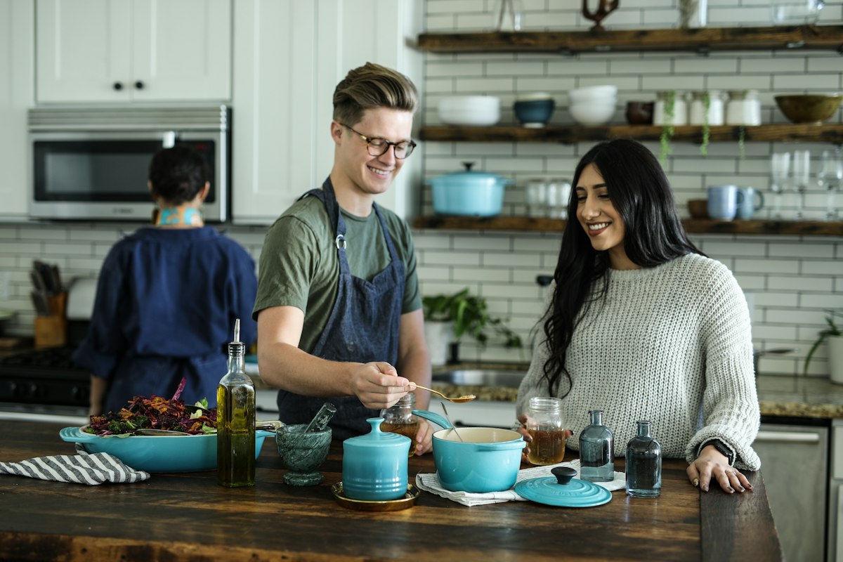 Two smiling people prepare food at a wooden kitchen island while a third person works behind them.