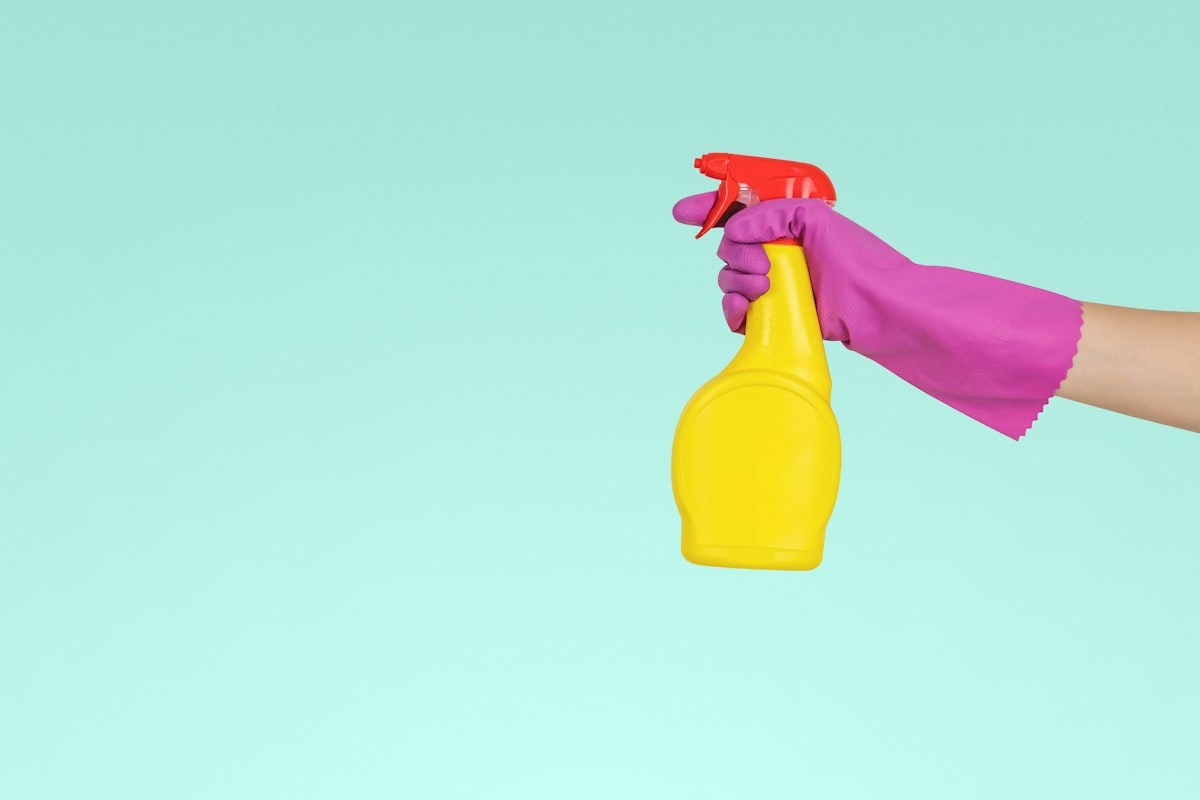A hand in a pink rubber glove holds a yellow spray bottle against a pale green background.